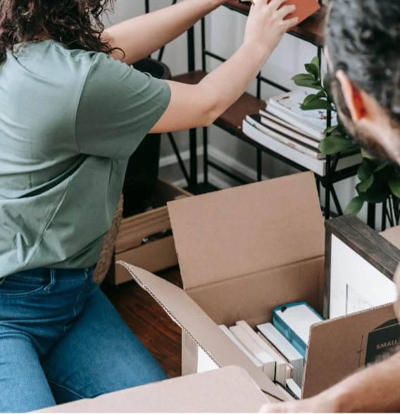 A person in a green shirt organizes items on a shelf while next to an open box containing books and assorted items.