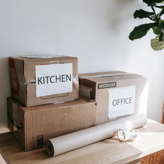 Three stacked boxes labeled "KITCHEN" and "OFFICE" on a wooden surface, with a roll of paper and tape nearby. Green plant in the background.
