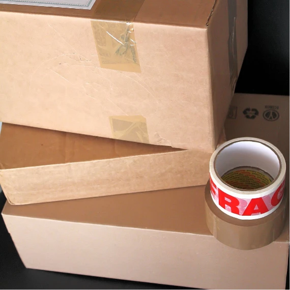 Three stacked cardboard boxes with adhesive tape, including a roll of "FRAGILE" tape, against a dark background.