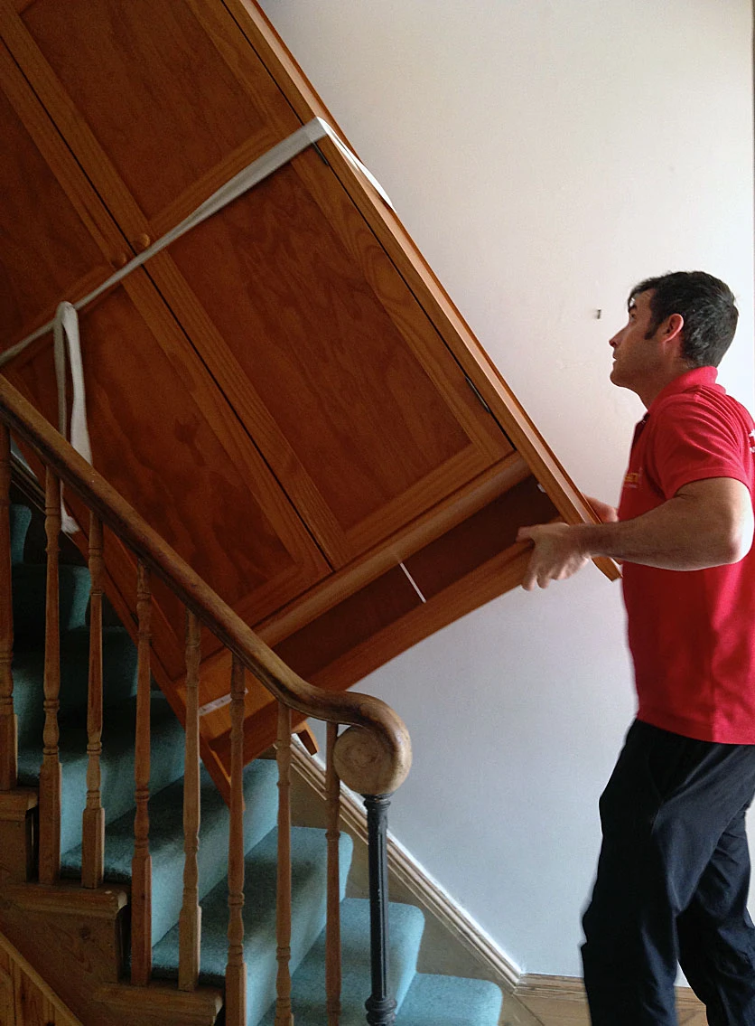 A person in a red shirt lifts a large wooden cabinet up a staircase, using a strap for support. Wooden banister and carpet visible.