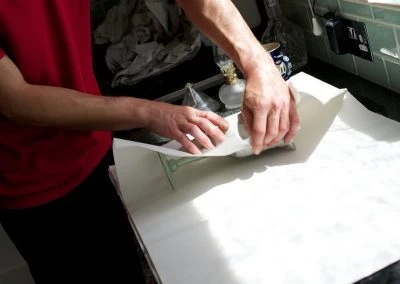 A person in a red shirt carefully folds a large sheet of paper on a kitchen countertop, with natural light illuminating the scene.