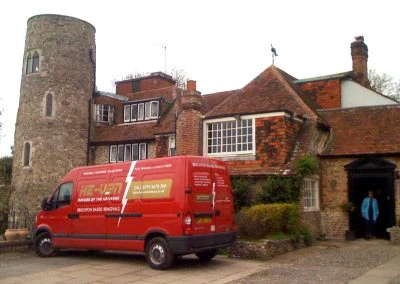 A red delivery van is parked in front of a stone tower house, showcasing a mix of traditional and modern architectural styles.
