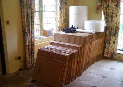 A stack of cardboard boxes with packing materials is arranged in a room with floral curtains and large windows.