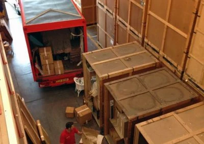 View from above of a red delivery truck unloading boxes in a storage facility with wooden crates and a worker organizing items.
