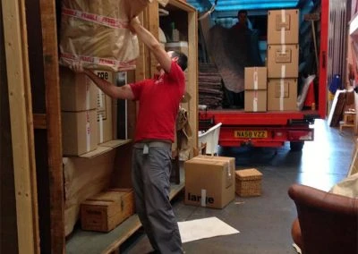 A person in a red shirt is lifting a package inside a storage area filled with boxes and furniture. A truck is visible in the background.