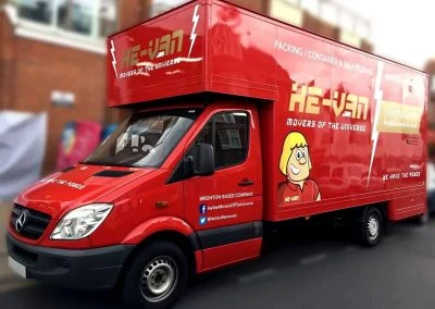 Red moving truck with cartoon character and text "He-Van Movers of the Universe." Displays service details on side.