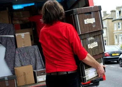 Person in a red shirt carries stacked boxes while unloading a moving van. Moving supplies and a residential area are visible in the background.